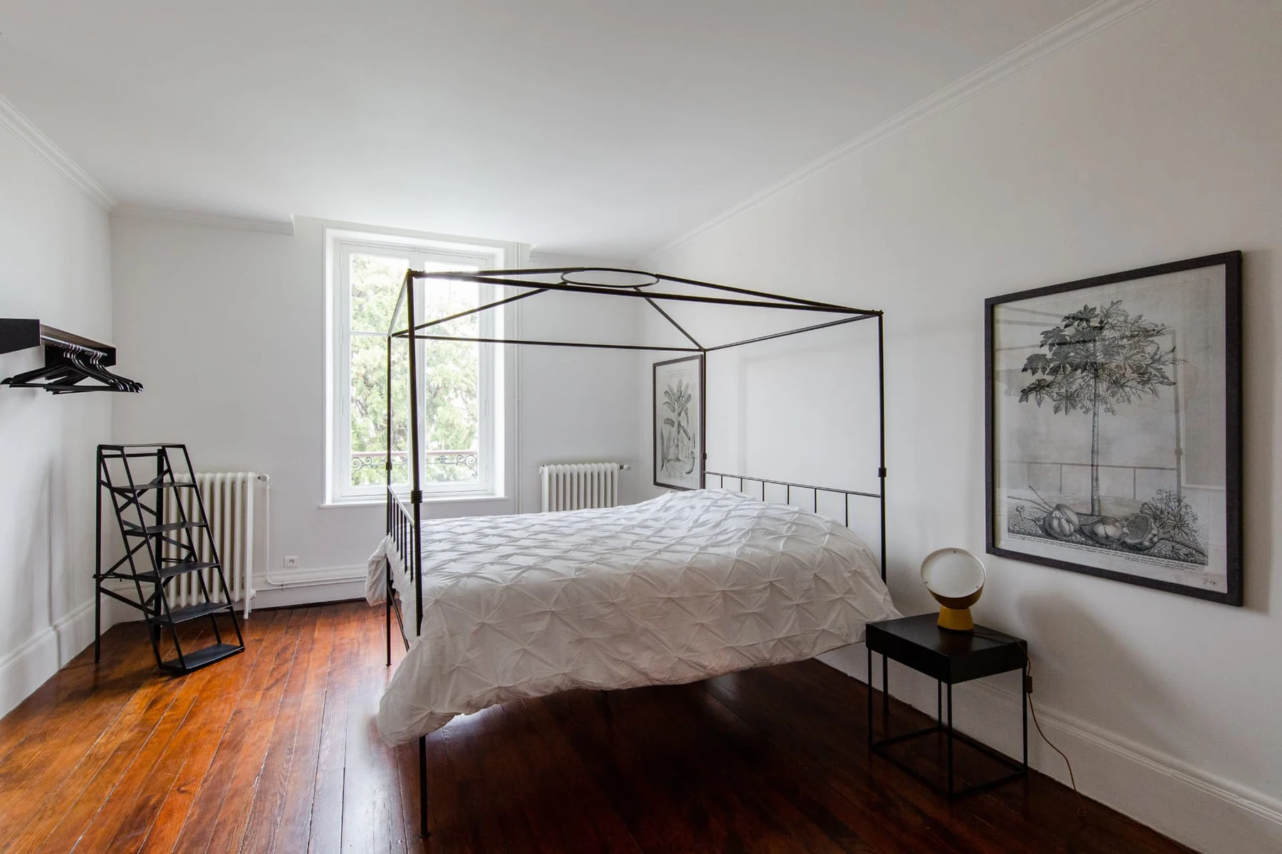 Chambre 1, lit à baldaquin en ferronnerie noire, murs blancs et haute fenêtre ouverte sur les arbres