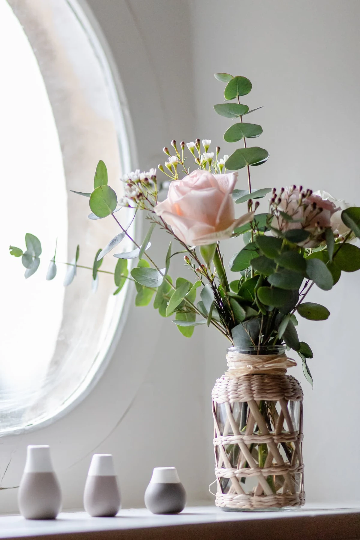 Bouquet de roses et d'eucalyptus dans un vase tressé devant une fenêtre ovale