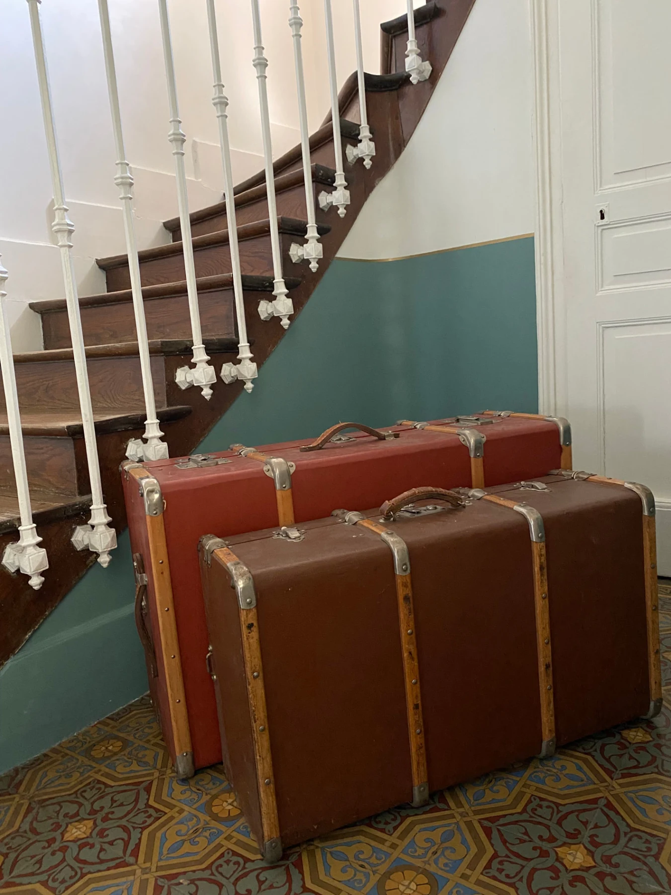 Deux valises anciennes posées sur les carreaux de ciment de l’entrée, au pied de l’escalier en bois