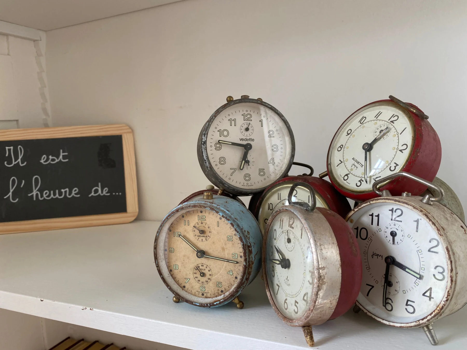 Collection de réveils anciens sur une étagère blanche, à côté d’une ardoise d’écolier