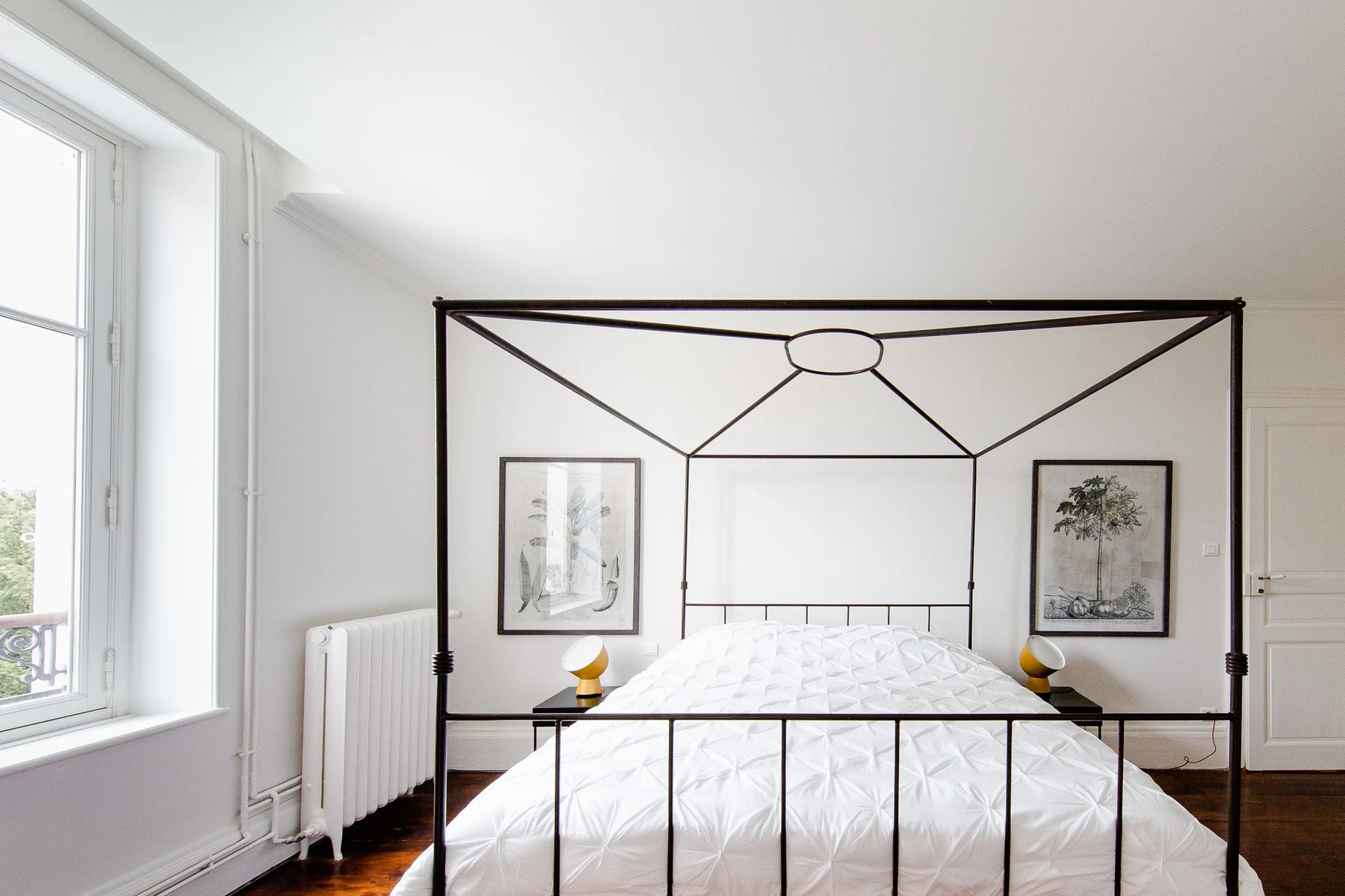 Le lit à baldaquin en fer noir sous le ciel blanc, lithographies botaniques et lampes jaunes