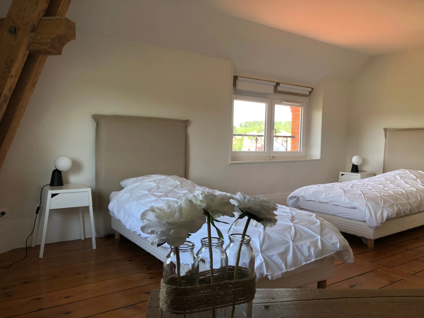 Chambre 7 : deux lits simples à tête de lit beige, chevets blancs et lampes globes, sous une poutre de bois