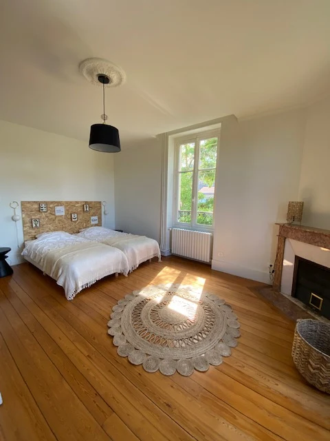 Chambre au tapis rond en jute, lit blanc, cheminée en marbre rose et fenêtre sur les arbres