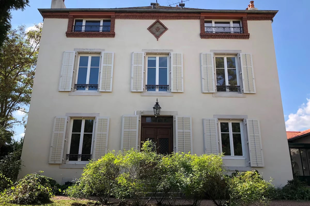 Façade de la Maison 1881 un jour de soleil, volets blancs ouverts et rosiers en fleurs