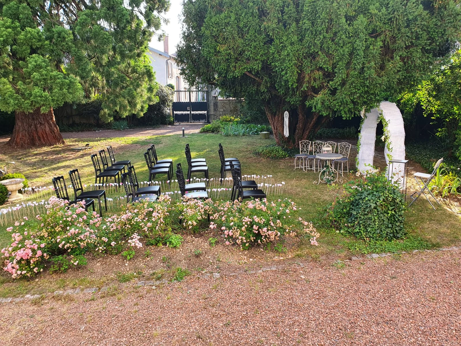 Cérémonie laïque dressée dans le jardin : chaises noires, arche blanche et rosiers en fleurs