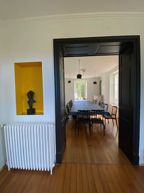 La salle à manger vue à travers les portes noires : longue table et chaises, niche jaune avec buste